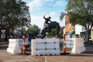 The downtown Stillwater roundabout at 10th Avenue and Main Street surrounded by jersey barriers and traffic cones, with the bronze bronco buster statue visible above the bare island. 