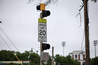 A school zone speed limit sign reading 20 mph when flashing on a Stillwater street, with stadium lights visible in the background.