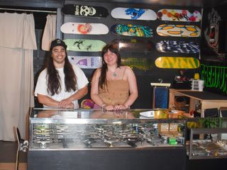Ivan Serrano, left, and Isa Edge smile behind the glass display counter at i&i Skateshop in Stillwater, with skate decks mounted on the wall behind them.