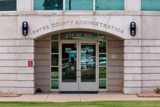 North entrance of the Payne County Administration Building at 315 W. 6th Ave. in Stillwater, Oklahoma, showing glass double doors under an arched stone facade.