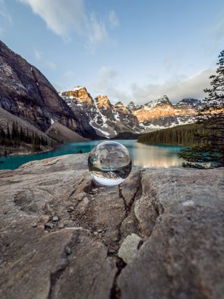 A crystal lens ball on a rock reflects turquoise water and snow-capped mountains at Moraine Lake in Banff National Park, taken by photographer Justin Eubanks.