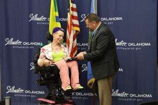 Mayor Will Joyce hands Allie Williams, who uses a power wheelchair, a proclamation for Spinal Muscular Atrophy Awareness Month in front of a Stillwater, Oklahoma backdrop.