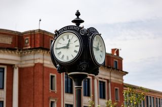 A four-faced black street clock lettered "Payne County" and "Stillwater" stands in front of the red brick Payne County Courthouse in downtown Stillwater.