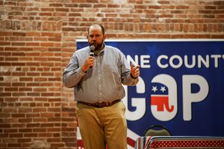 Dillon Travis speaks into a handheld microphone at a Payne County GOP candidate forum, a Payne County GOP banner and brick wall behind him.