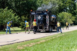 Five Scodeller Construction workers in safety vests seal cracks behind a steaming sealant truck on a tree-lined residential street in Stillwater on July 25, 2025.