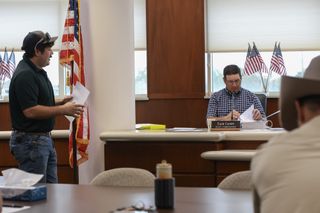 Pierce Taylor stands at left speaking to Payne County commissioners while Zach Cavett sits at the dais reviewing a thick binder of documents at the Sept. 21, 2026, meeting in Stillwater.