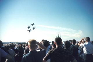 Four Air Force Thunderbirds F-100 jets fly in diamond formation trailing white smoke as a crowd, seen from behind, shades their eyes and watches from an airport tarmac in 1960.