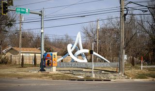 White looping sculpture Interlace above a Stillwater Oklahoma sign at Sixth Avenue and Western Road, with a 6th Ave/Highway 51 street sign and painted utility box nearby.