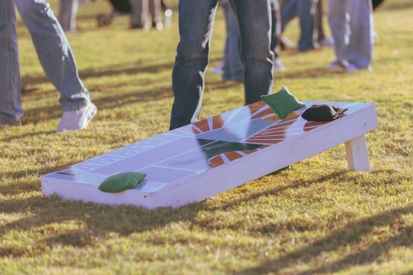 A close-up of a cornhole board on a grassy field with players standing nearby, taken during a sunny outdoor event in Stillwater.