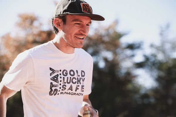 Stone Lambert wearing a white "Stay Gold Lucky Safe Run4Grady" t-shirt and cap, smiling while holding a small urn containing his brother's ashes during an outdoor run