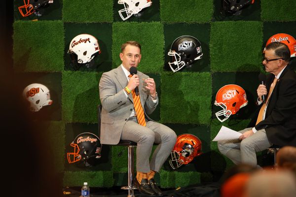 Eric Morris in gray suit with orange tie speaks into microphone during Oklahoma State football coach introduction, with OSU helmets displayed on wall behind him