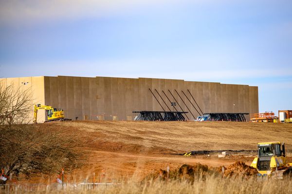 Large concrete tilt-up walls of Google's data center under construction, supported by black steel bracing, with excavators and bulldozers working on the graded dirt site under a blue sky.