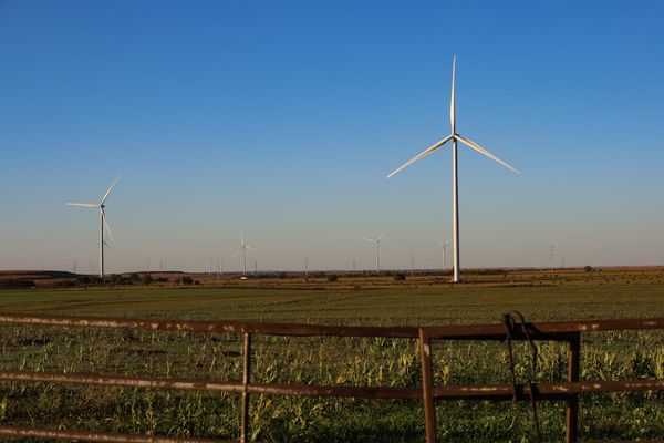Wind turbines rise above green agricultural fields with a rusty barbed wire fence in the foreground under a clear blue sky in Noble County, Oklahoma
