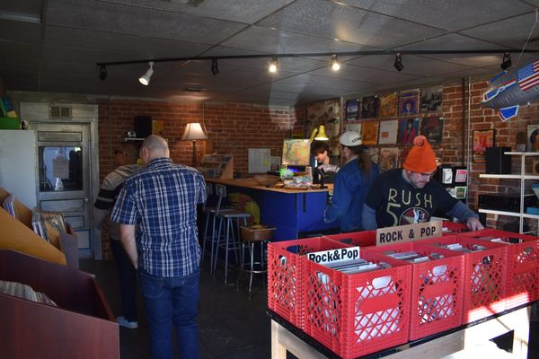 Record store interior with customers browsing vinyl in red crates in foreground. Owner at checkout counter in background. Brick walls with album posters, track lighting.