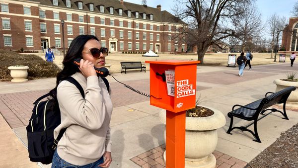 Student in sunglasses and backpack talks on orange OSU phone booth labeled "The Code Calls" on brick campus walkway with students passing in background