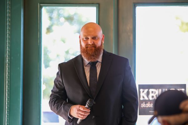 Luke Kruse wearing dark suit and tie holds microphone while speaking at indoor campaign event with turquoise door frames and campaign sign visible in background.