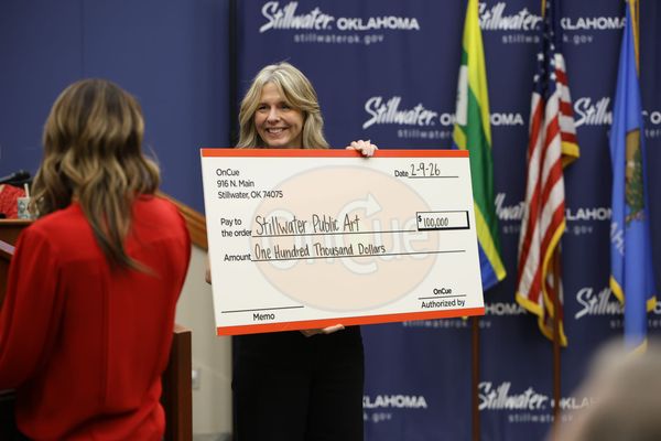 Marla Ruark holds ceremonial $100,000 check from OnCue to Stillwater Public Art at city council meeting with Stillwater, Oklahoma backdrop and flags