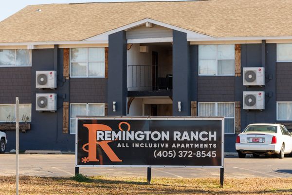 Sign reading "Remington Ranch All Inclusive Apartments" stands before weathered gray apartment building with AC units mounted on exterior walls.