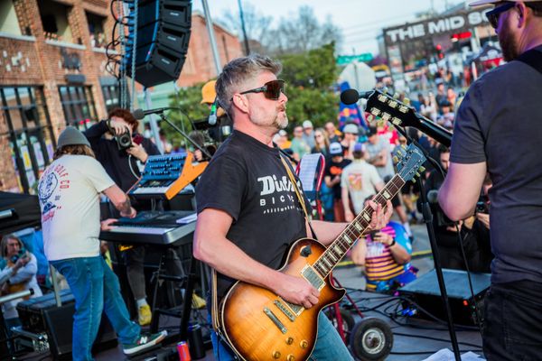 Bobby Wintle plays a sunburst Les Paul guitar at a microphone on an outdoor stage during The Mid South 2024, with a keyboard player behind him, a packed crowd.