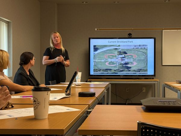 arolyn Walstad stands before a screen showing an aerial view of Strickland Park's four ball fields while presenting to seated Tourism Advisory Committee members.