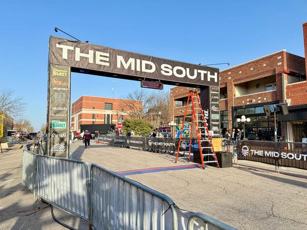 The Mid South finish line arch spans West 7th Avenue in downtown Stillwater during setup, with sponsor banners, crowd barriers, and a timer display visible under clear blue skies. 