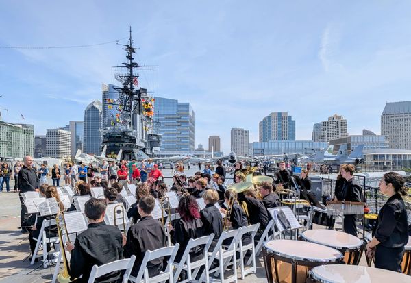 A high school concert band in black uniforms performs on the USS Midway flight deck, surrounded by vintage aircraft and the San Diego skyline.