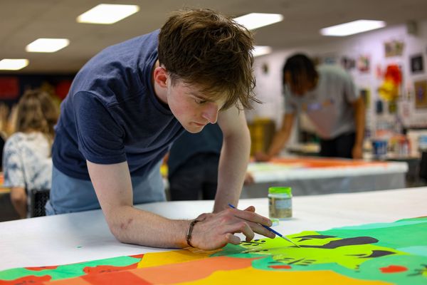 OSU student Jake Warner leans over a large fabric panel, painting with a fine brush in a studio classroom at the Prairie Arts Center, as classmates work in the background.