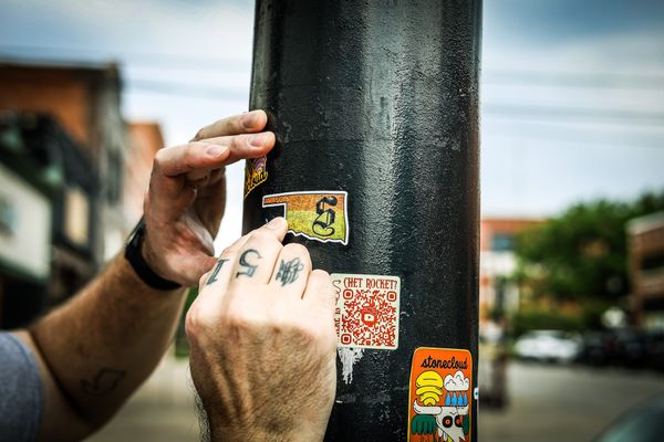 Close-up of hands pressing an Oklahoma-shaped sticker with The Stillwegian's blackletter S onto a sticker-covered black pole in downtown Stillwater, surrounded by other local stickers