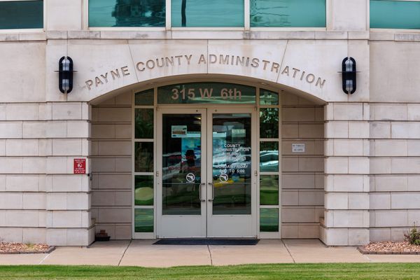 North entrance of the Payne County Administration Building at 315 W. 6th Ave. in Stillwater, Oklahoma, showing glass double doors under an arched stone facade.