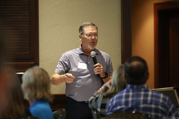 Chuck Hall speaks into a handheld microphone before a seated audience at a Stillwater Chamber of Commerce candidate luncheon.