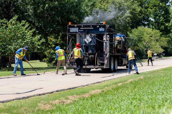 Five Scodeller Construction workers in safety vests seal cracks behind a steaming sealant truck on a tree-lined residential street in Stillwater on July 25, 2025.