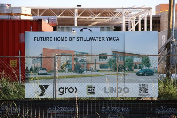 A construction sign showing a rendering of the new Stillwater YMCA stands behind a chain-link fence, with steel framing of the building under construction visible in the background.