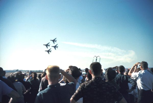Four Air Force Thunderbirds F-100 jets fly in diamond formation trailing white smoke as a crowd, seen from behind, shades their eyes and watches from an airport tarmac in 1960.