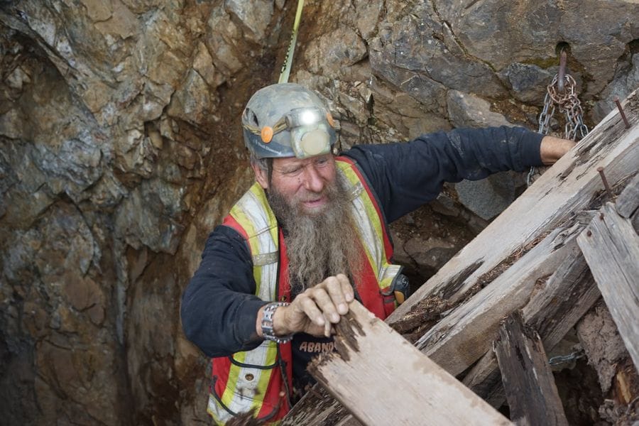 Frank Schlichting standing on the collapsed timbers of an ore bin high above a valley, snowy mountain behind.
