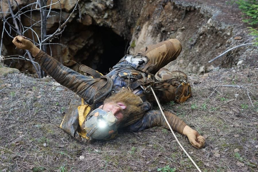 Frank Schlichting emerging by headlamp from a small mine portal nearly closed over by moss, roots and fallen earth.