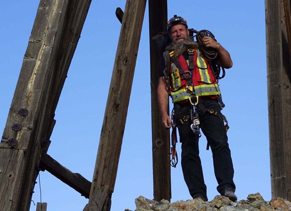 Frank Schlichting in full climbing kit with a coiled rope over his shoulder, standing among old mine timbers against a blue sky in BC.
