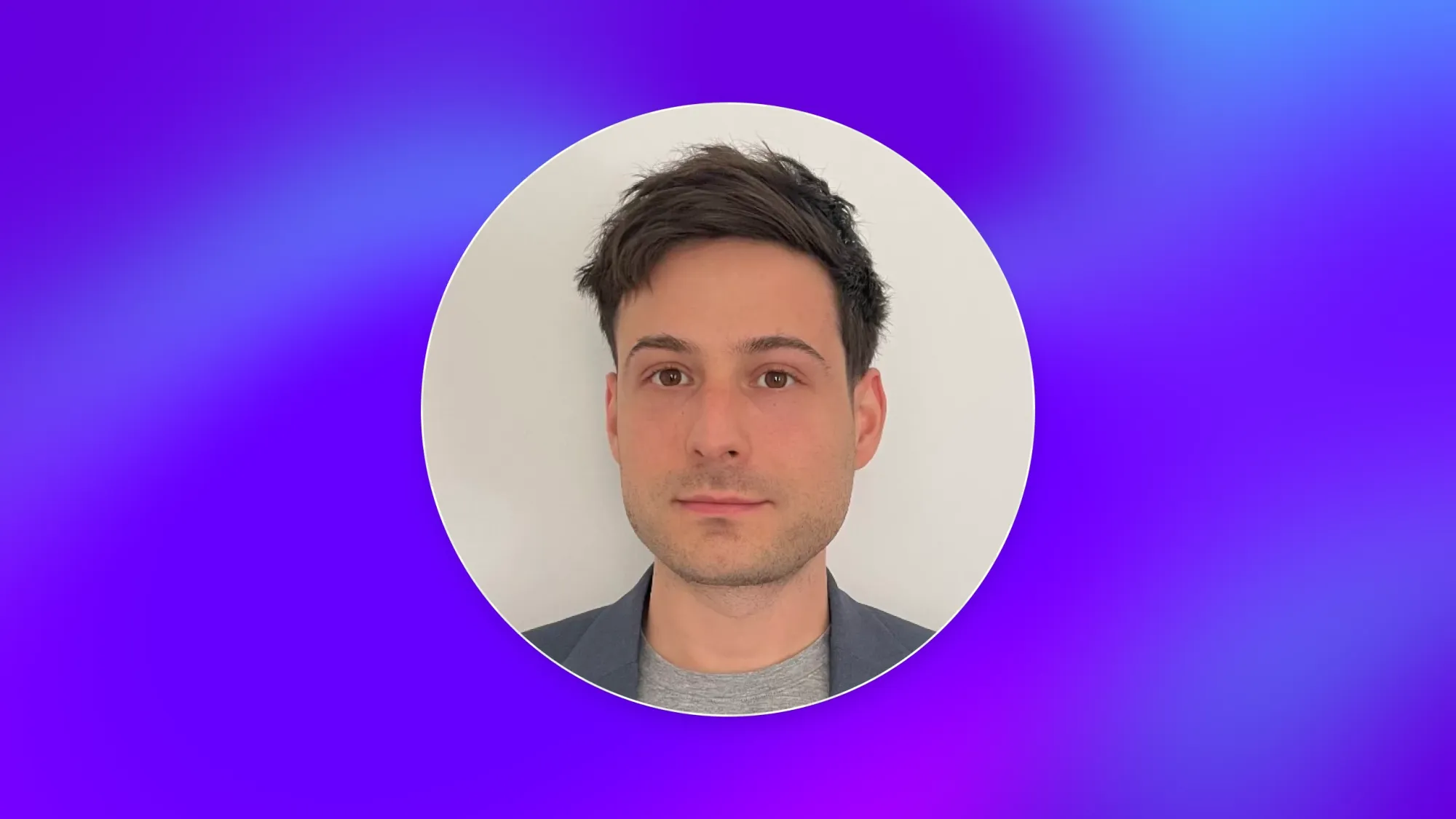 Professional headshot of a man with short dark hair wearing a blazer and gray shirt on a purple-blue gradient background