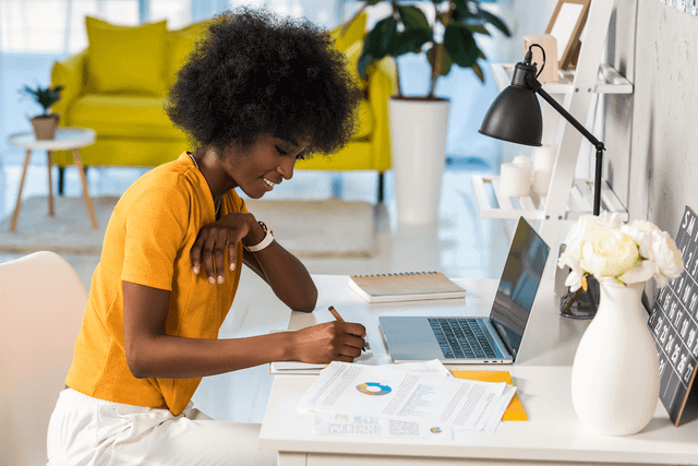 Woman in yellow blouse sitting at desktop in front of laptop.