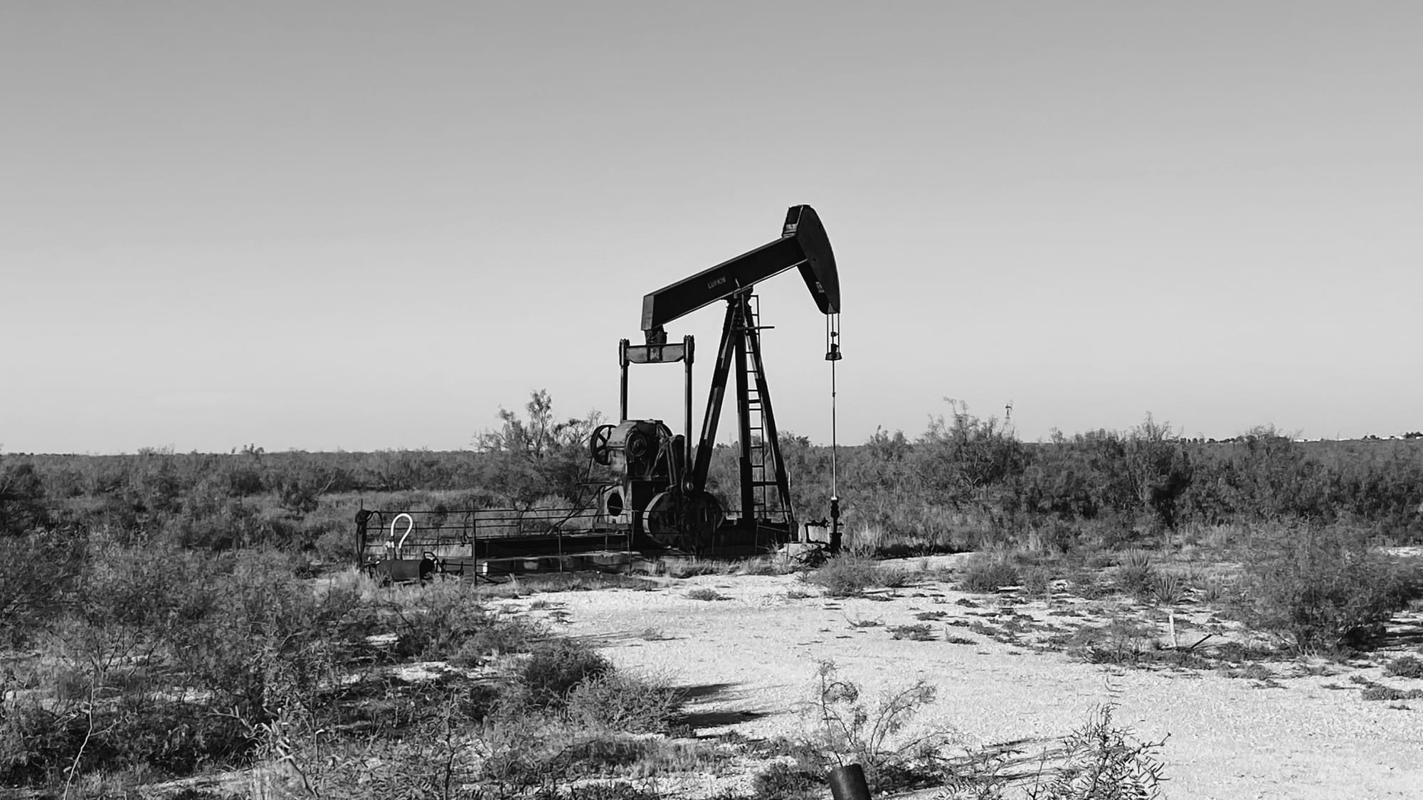 Black & white photo of a desert landscape with a "nodding donkey" or lone pump jack - a machine for pumping oil up from underground wells 