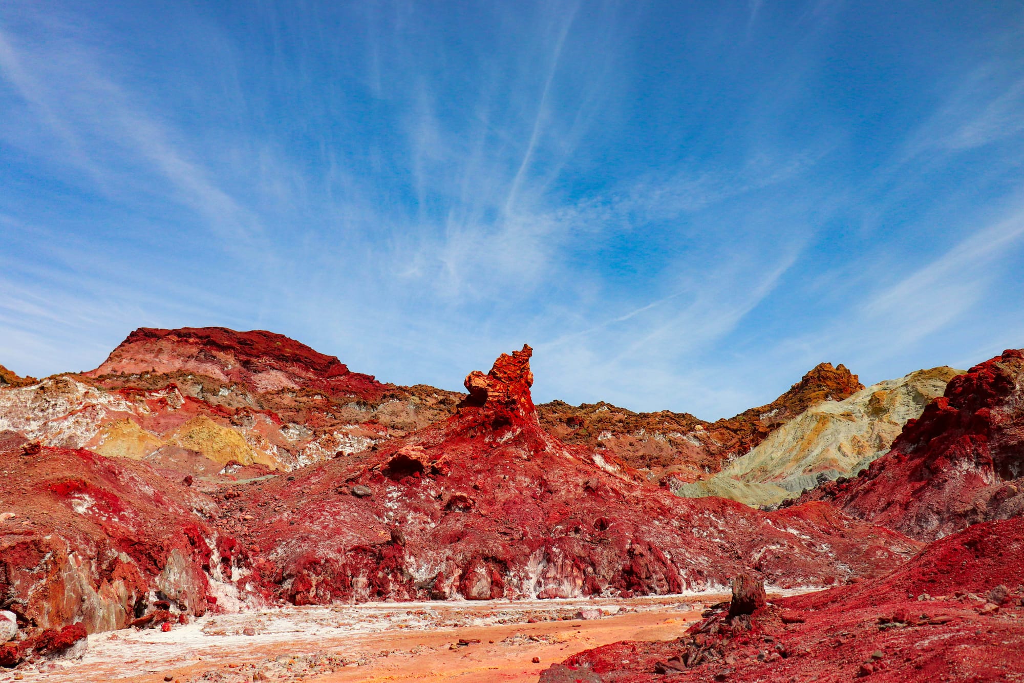 Bright red rock formations on Hormuz Island beneath a deep blue sky with wispy white clouds.
