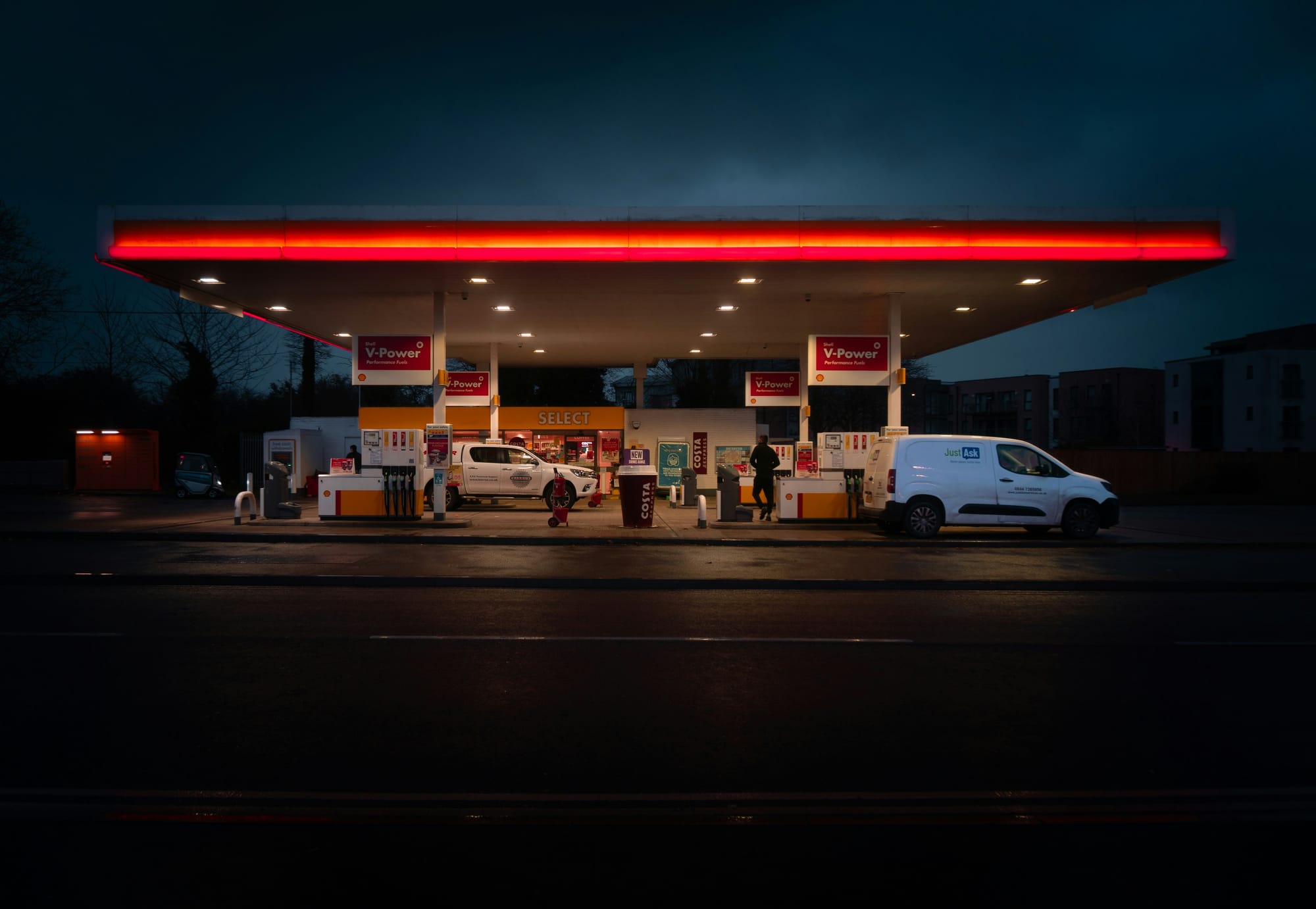 Mid-distance photo of a petrol station, probably in the UK, with a couple of white vehicles, at night