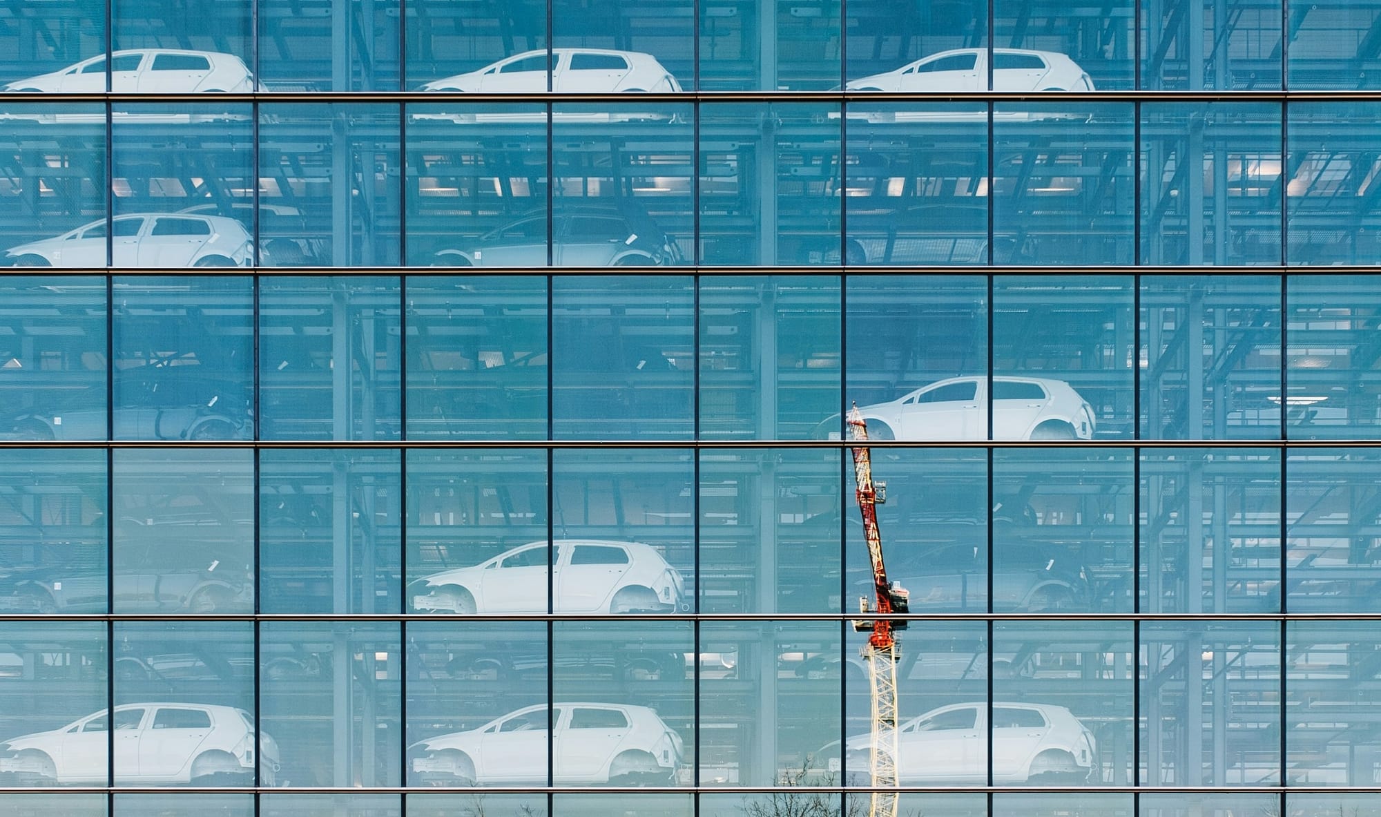 Exterior of a glass-walled building, multiple floors each containing multiple identifical white, black and silver compact cars. Volkswagen factory in Dresden. 