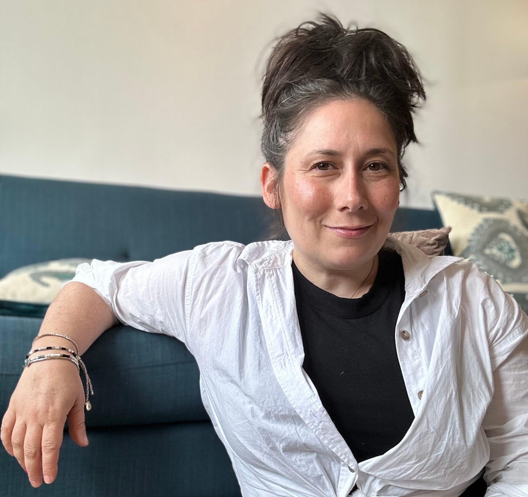 Headshot of today's guest, Zoë Bisbing, siting on the floor with her arm leaning on a blue sofa, wearing a black t-shirt and white over-shirt