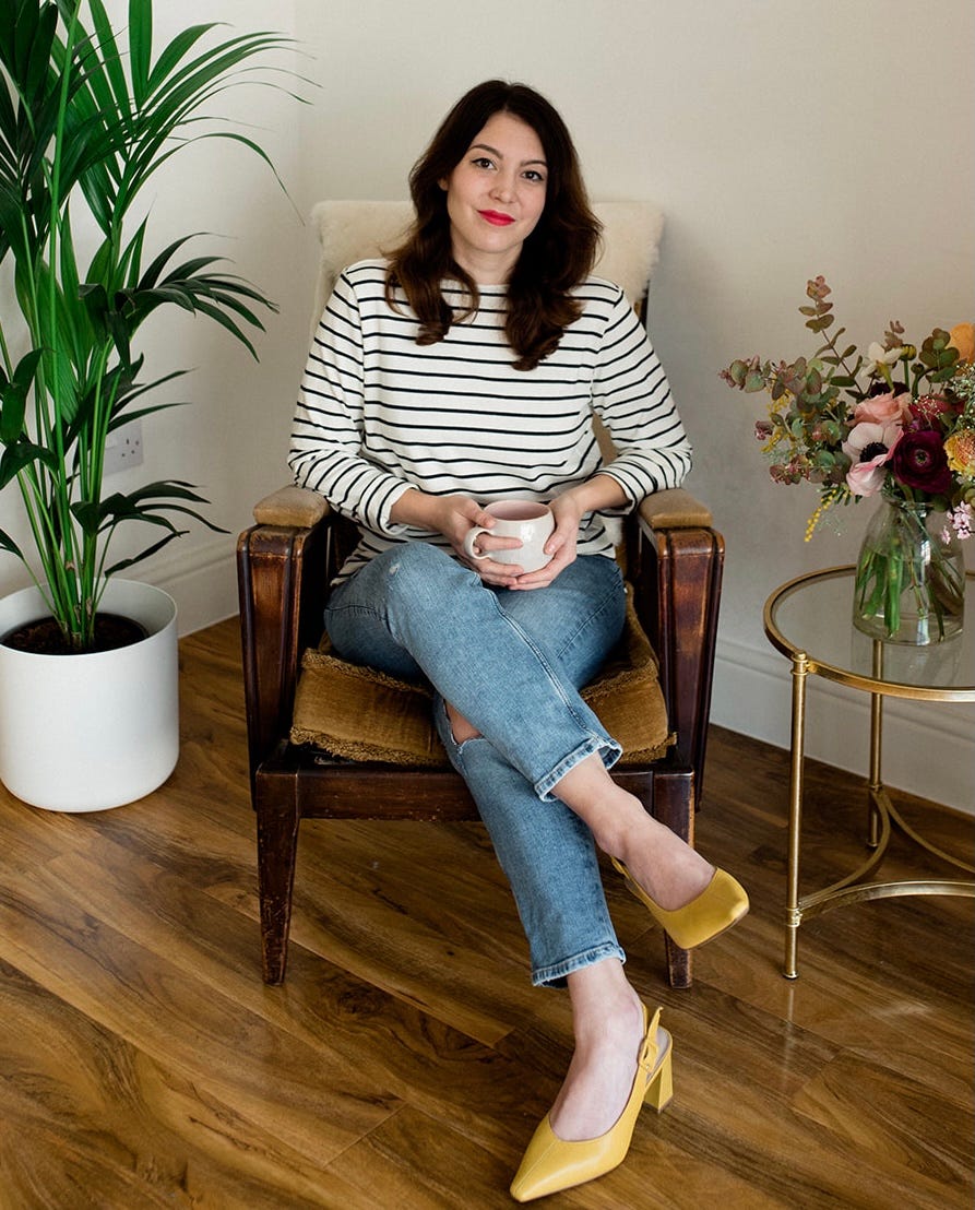 Today's guest, Jennie Agg, sitting in an armchar, holding a cup, and surrounded my plants and flowers against a plain white wall.