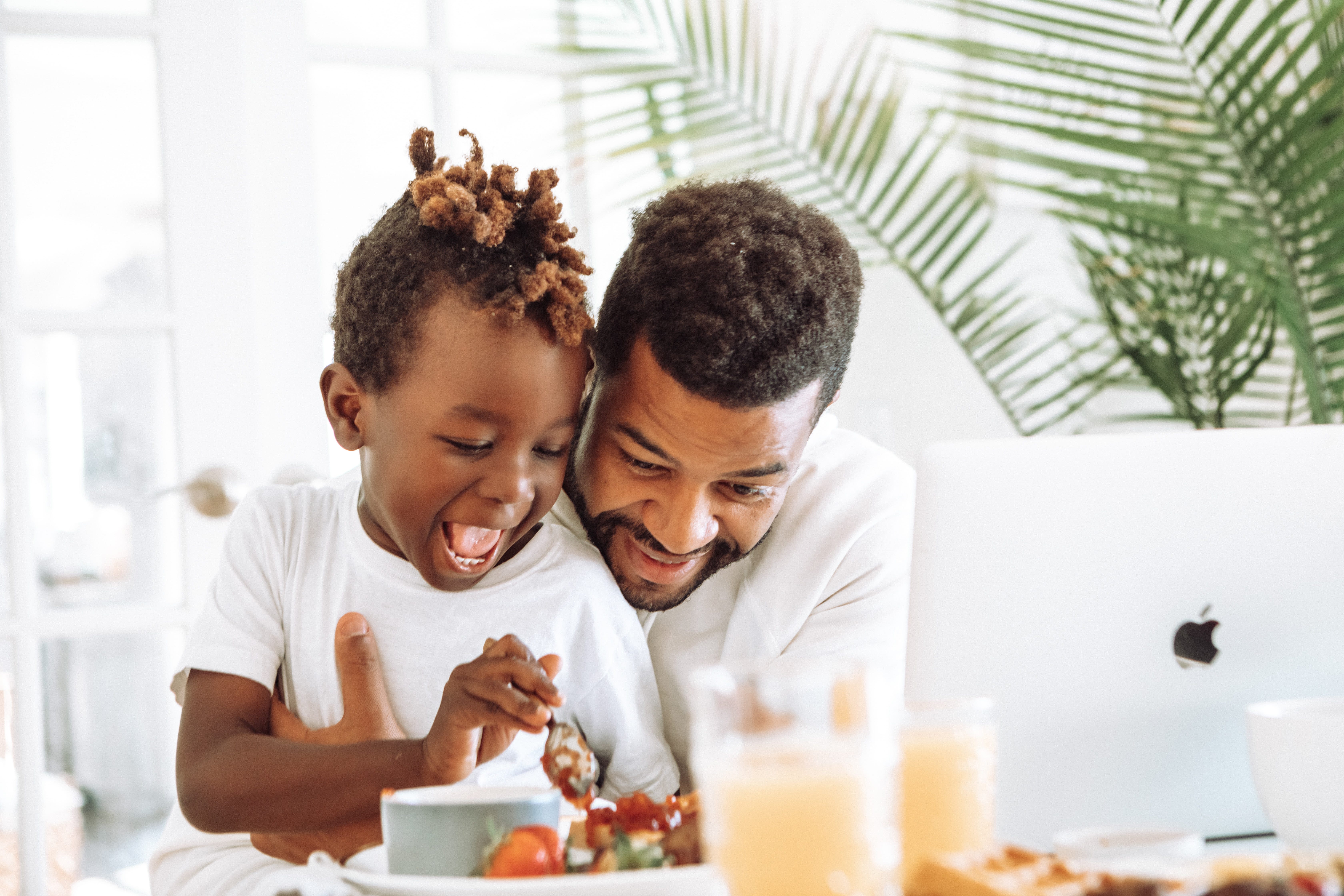 A man and child in white crew neck t-shirts, sat eating from white ceramic plate of food white an Apple Macbook in front of them