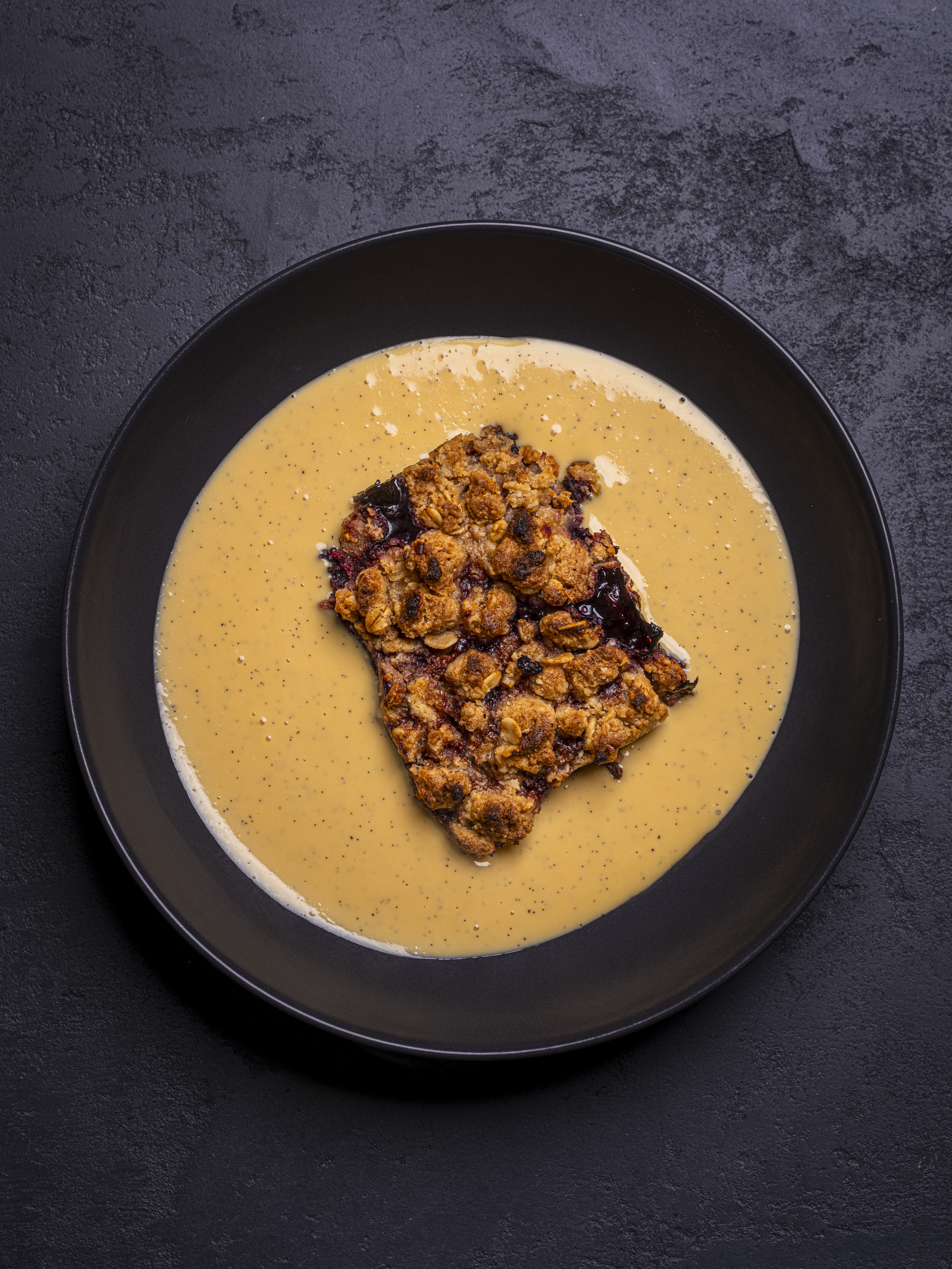 Overhead shot of Wild Blackberry Crumble with Real Vanilla Custard in a black bowl on a black background