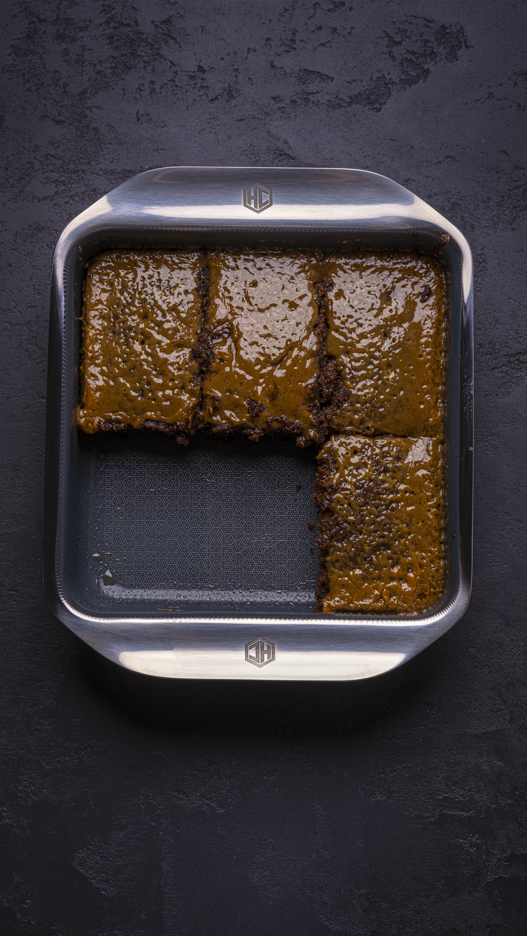 Overhead shot of sticky toffee pudding in a hexclad bakeware tray. With two pieces of the sticky toffee pudding missing on a dark background 