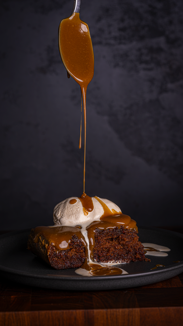 Sticky toffee pudding with vanilla ice cream and sticky toffee sauce being poured over the top from a spoon, on a black background and black plate 