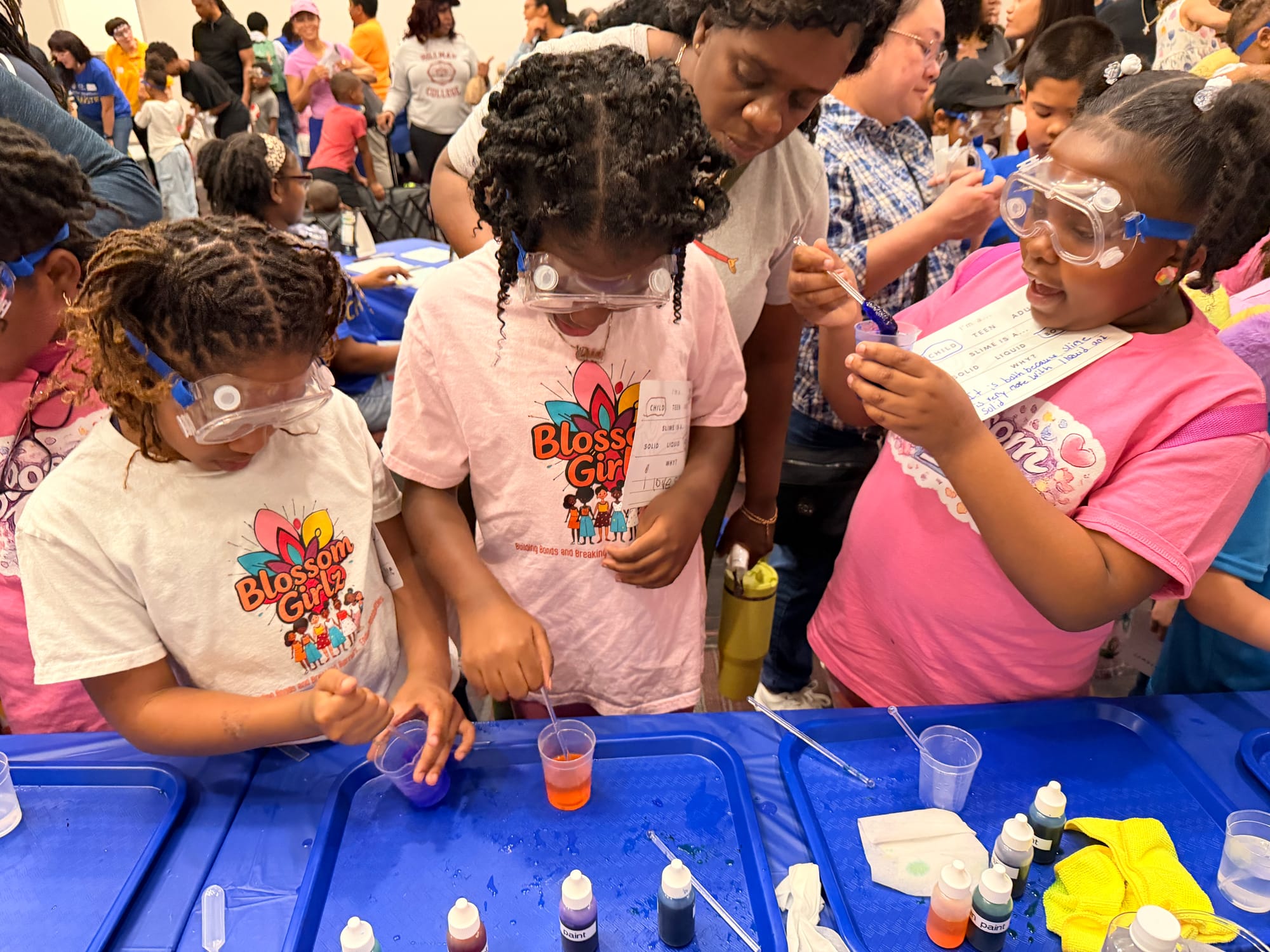 Several young girls wearing safety goggles and pink "Blossom Girlz" shirts enthusiastically mix colorful slime solutions in plastic cups at a science outreach table, guided by volunteers.