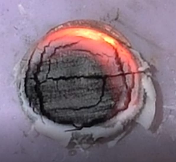 Close-up of a round, charred ember glowing orange along one edge as it smolders on a pale surface in a wind tunnel.
